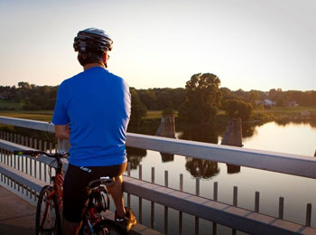 Homme à vélo vu de dos; il semble de tenir sur un pont au-dessus de l’eau.