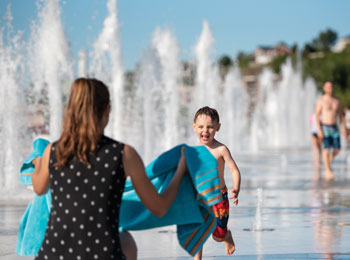 Jeune garçon dans des jeux d'eau en train de courir joyeusement vers sa mère, qui tient une serviette.